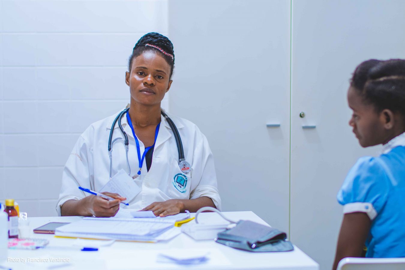 A woman in a medical coat with a stethoscope around her neck is taking notes of a young girl's medical record and looks into the camera