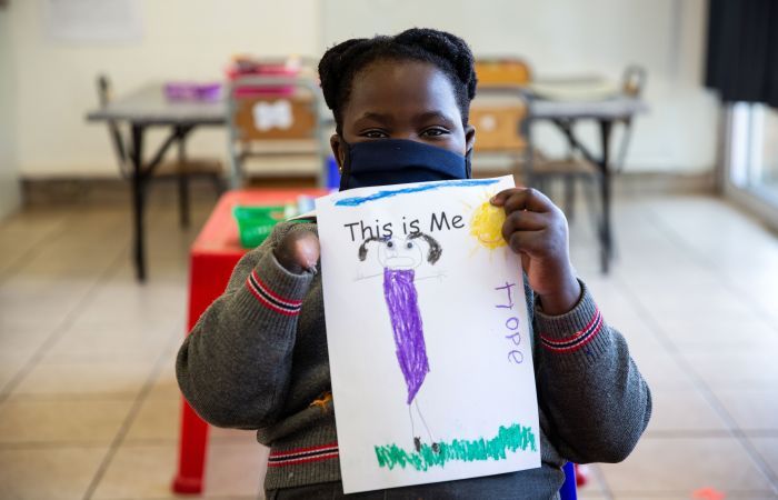 Girl whose right hand is missing holds up a drawing of herself entitled 'This is me' and her name, Hope, is visible on the side of the paper