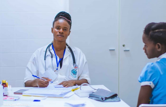 A woman in a medical coat with a stethoscope around her neck is taking notes of a young girl's medical record and looks into the camera